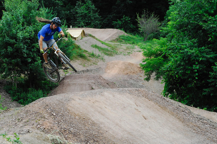 A mountain biker in a blue shirt and helmet performs a jump over a dirt ramp in a wooded area, surrounded by green trees and a well-maintained trail designed for cycling.