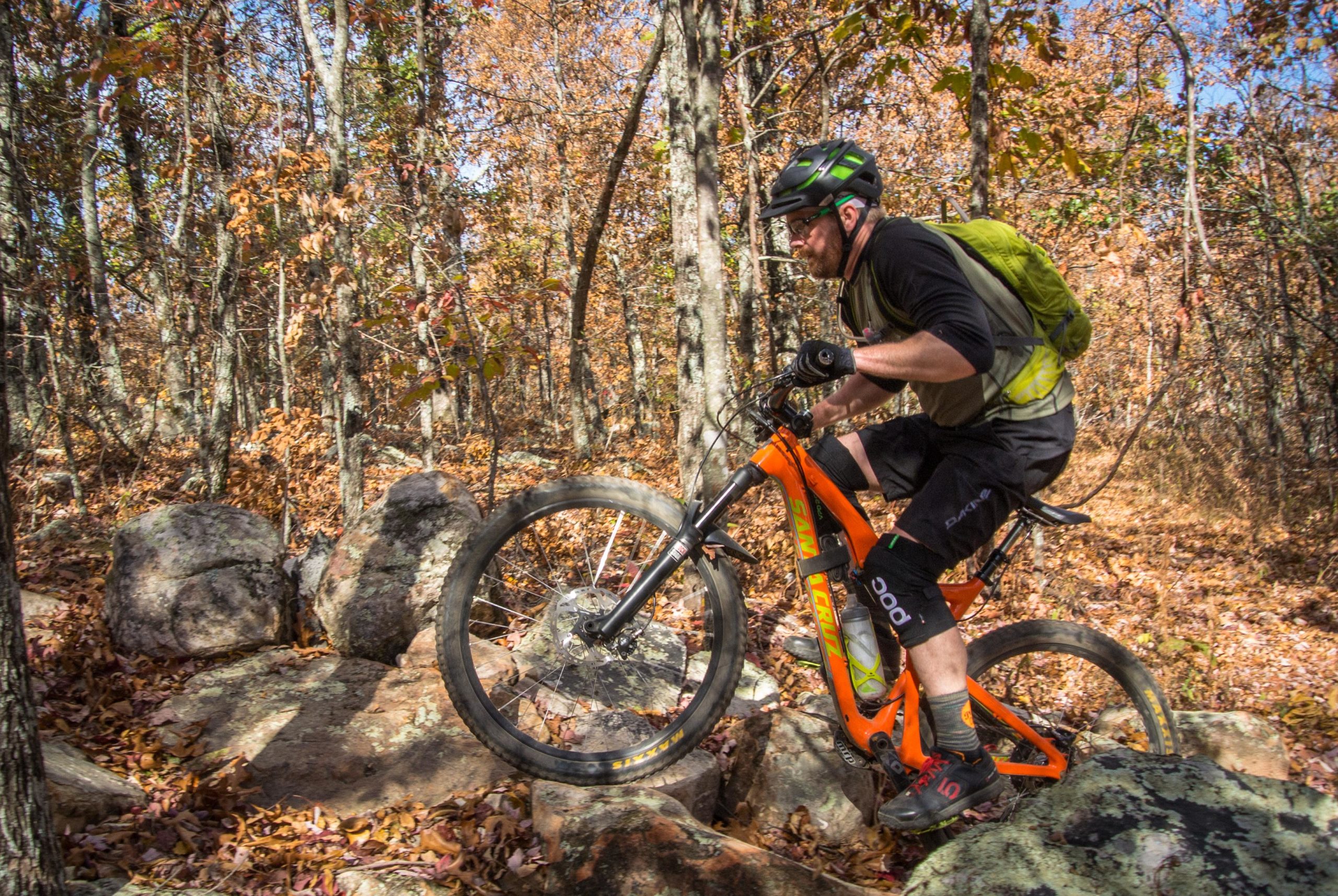A mountain biker navigating rocky terrain in a forest during autumn, surrounded by trees with orange and brown leaves. The biker is wearing a helmet and a backpack, focusing intently as they maneuver over the rocks. Coldwater Mountain mountain bike trail.