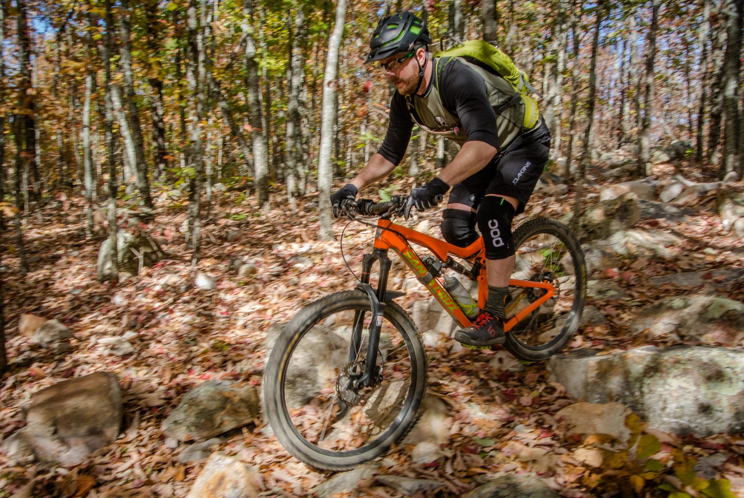 A mountain biker navigating a rocky trail surrounded by autumn foliage. The cyclist is wearing a helmet and protective gear, and their bike is prominently orange with black accents. The scene captures the motion and excitement of off-road biking amidst colorful leaves and trees. Coldwater Mountain mountain bike trail.