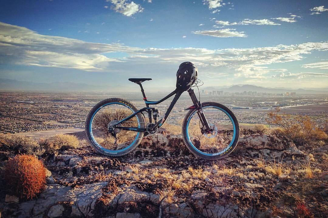 Giant Trance 27.5 2: A mountain bike with a helmet resting on the handlebars is positioned on rocky terrain, with a panoramic view of a valley and city in the background under a colorful sky.