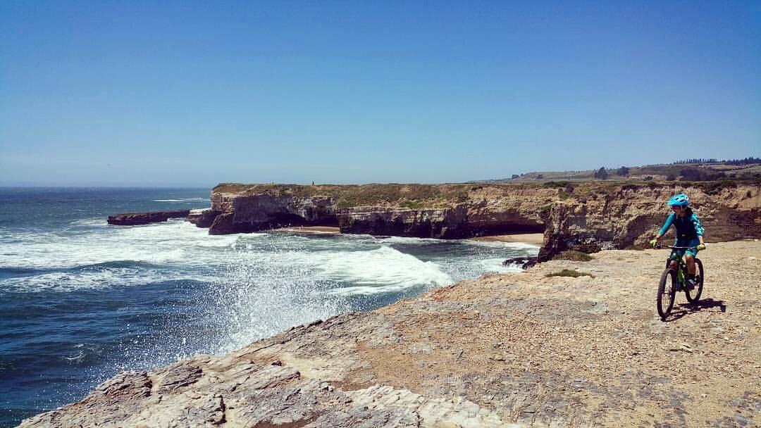 A child riding a bicycle on a rocky coastline, with waves crashing against the shore and a clear blue sky overhead. In the background, rugged cliffs and greenery are visible, along with a beach area. Wilder Ranch State Park mountain bike trail.