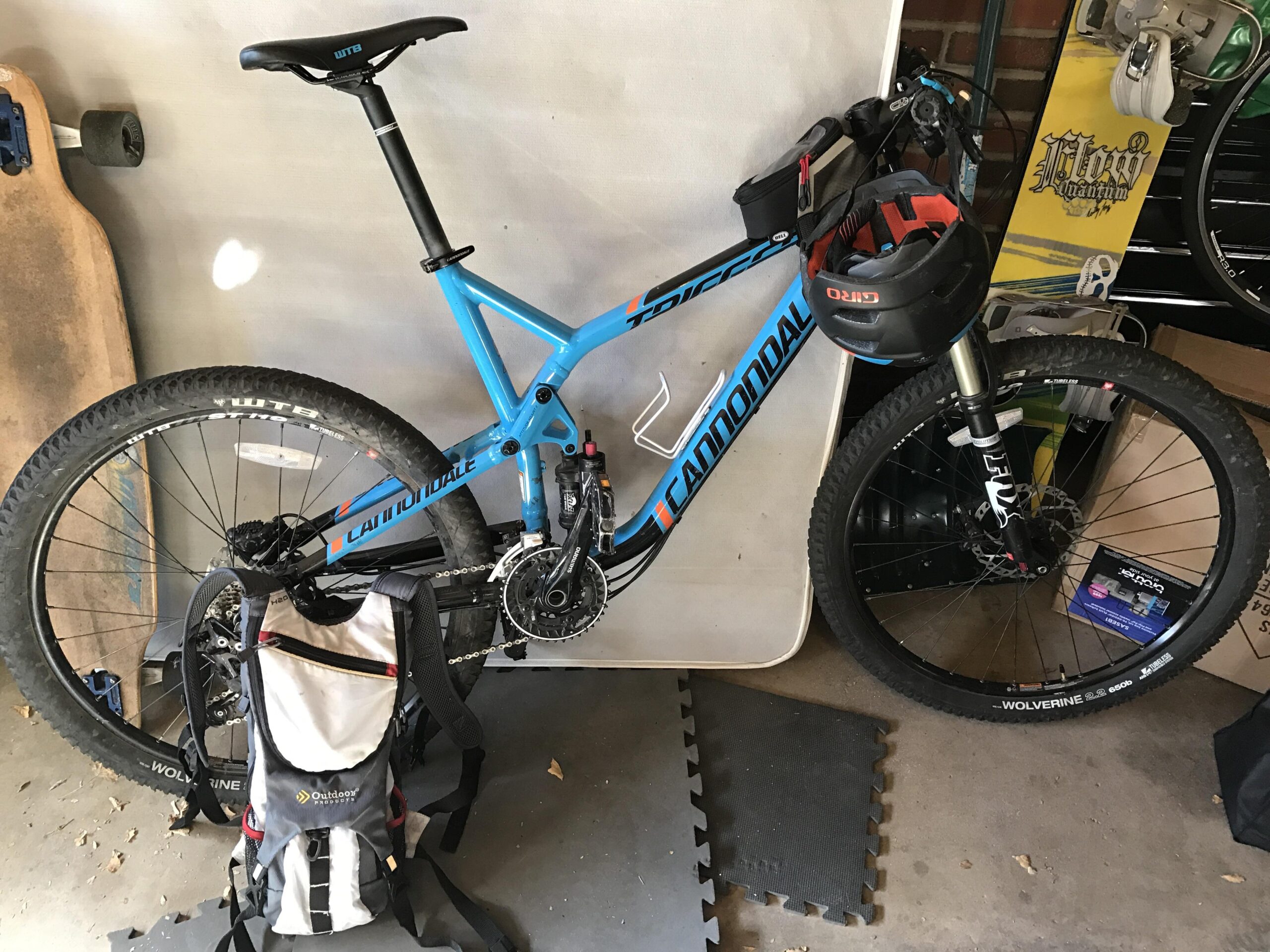 Cannondale Trigger 27.5 3: A blue Cannondale mountain bike leaning against a wall in a garage, equipped with a helmet mounted on the handlebars. A hydration backpack is positioned on the floor beside the bike, with skateboards and storage boxes visible in the background.