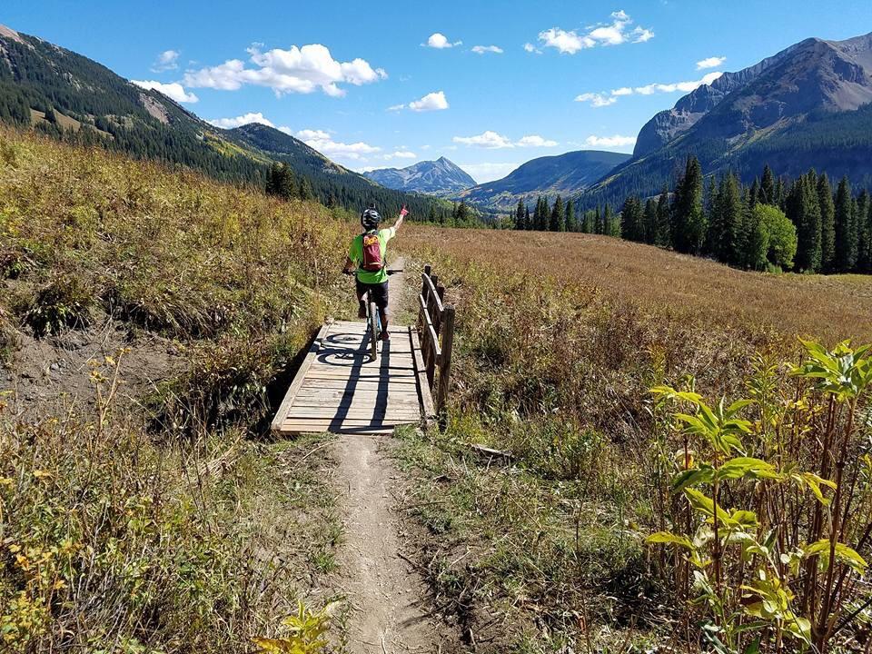A cyclist on a mountain bike stands on a wooden bridge in a scenic landscape, raising one hand toward the mountains in the distance. The area features lush greenery, tall grasses, and colorful autumn foliage under a bright blue sky with fluffy white clouds. Trail 401 mountain bike trail.