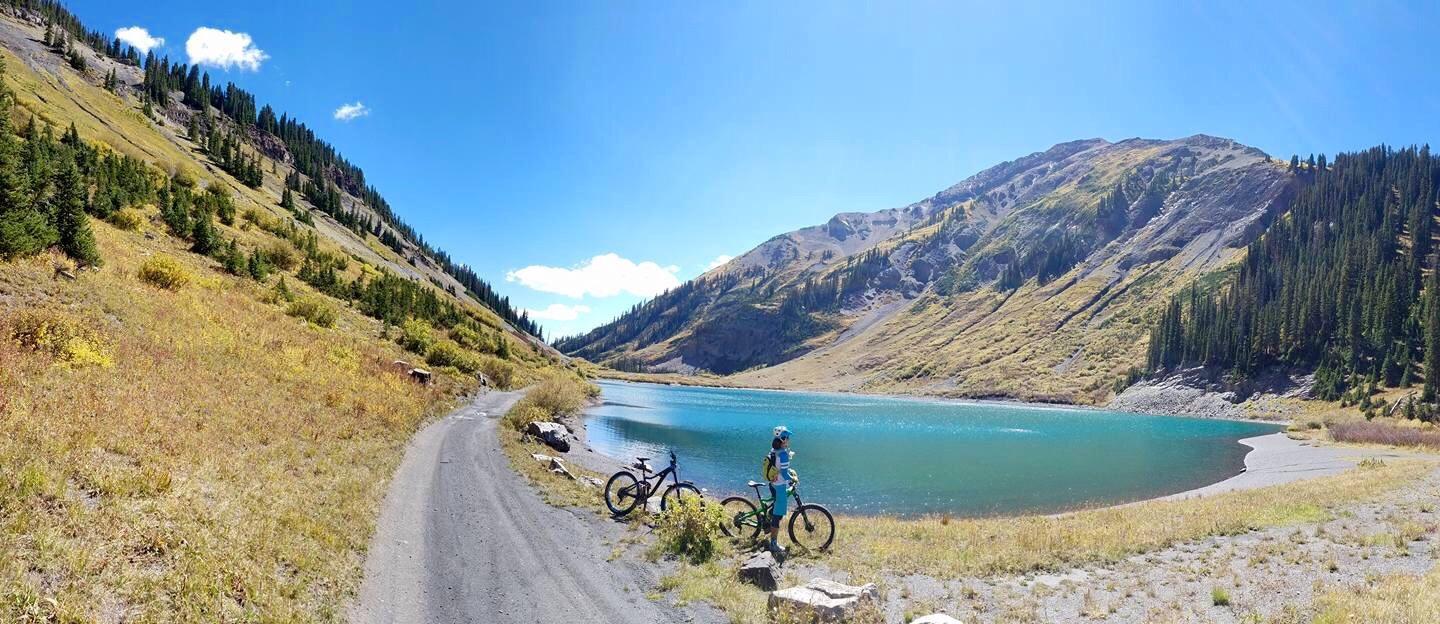 A scenic view of a mountain lake surrounded by hills and pine trees, with a person standing by the water's edge and two bicycles parked nearby. The sky is clear and blue, and the landscape features autumn colors in the foliage. Trail 401 mountain bike trail.