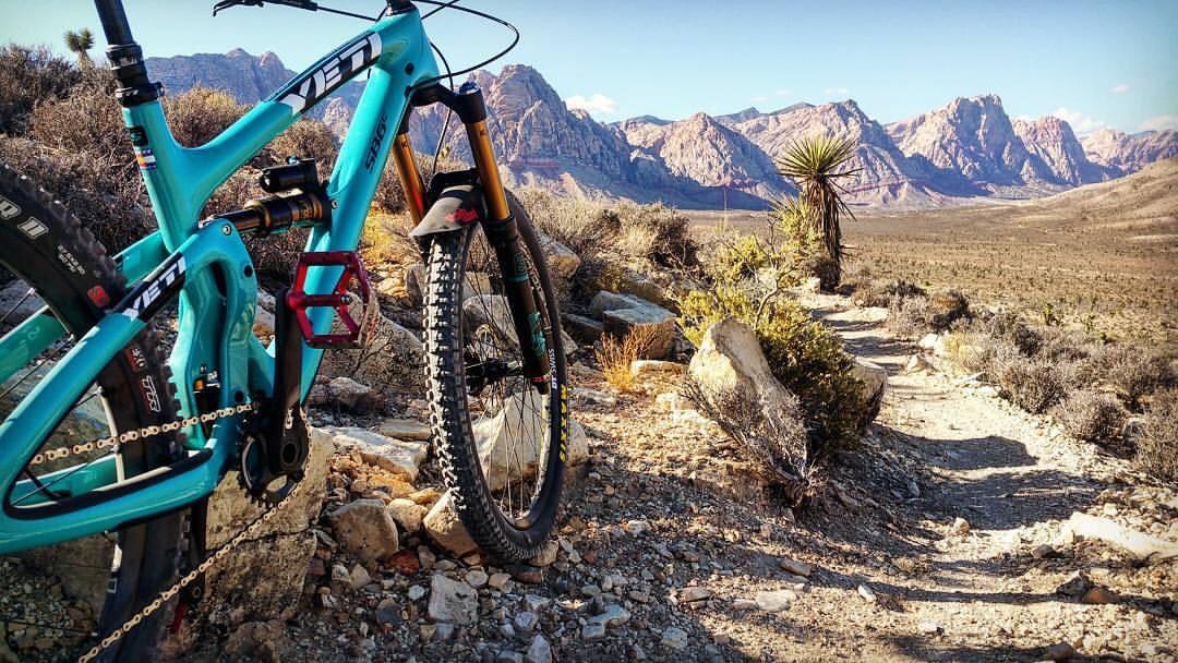 Yeti SB6: A turquoise mountain bike is parked on a rocky trail with a scenic desert landscape in the background, featuring rugged mountains and sparse vegetation. The sun is shining, creating a bright and vibrant atmosphere.