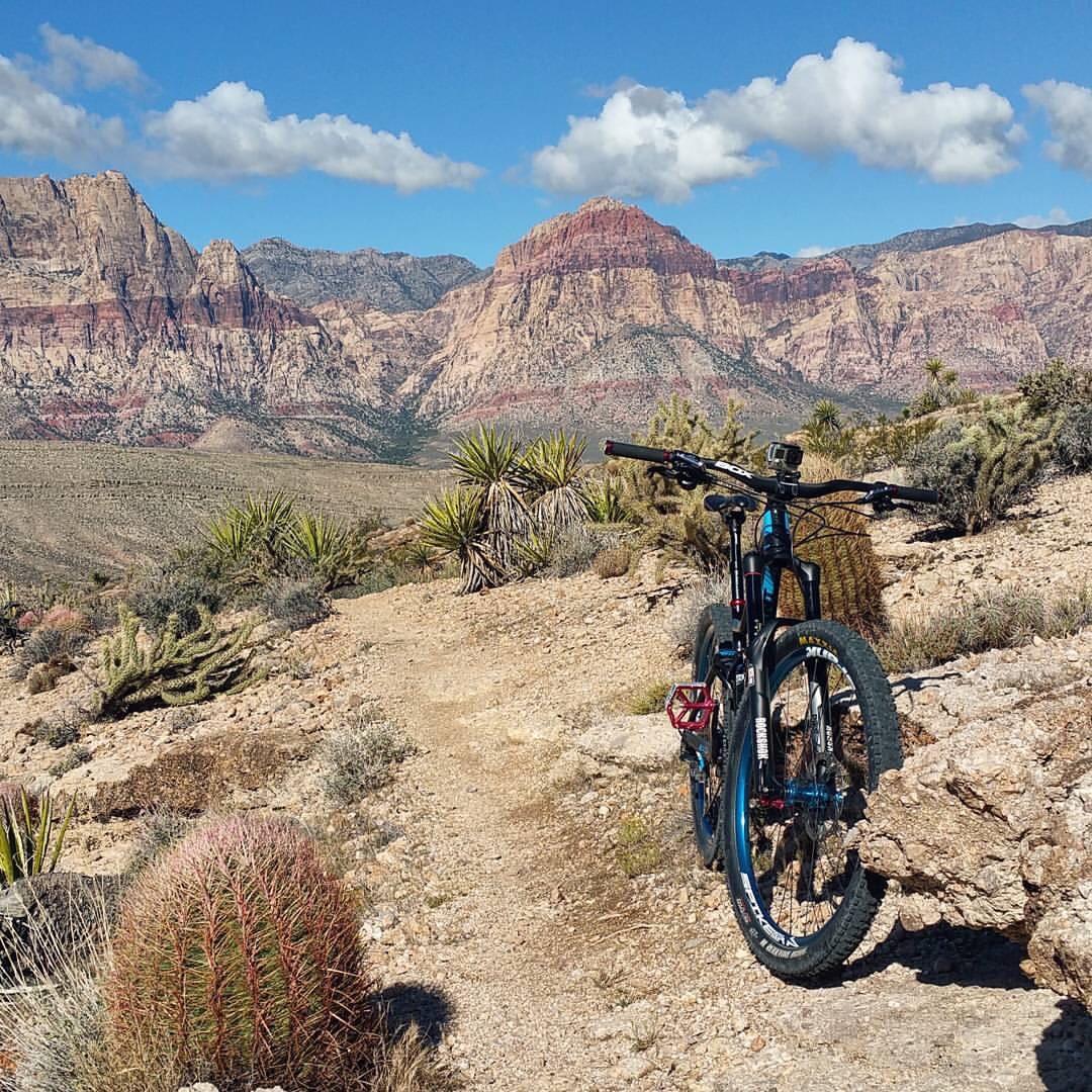 A mountain bike resting on a rocky trail, surrounded by desert vegetation, with rugged mountains and a blue sky featuring scattered clouds in the background. Cowboy Trails mountain bike trail.