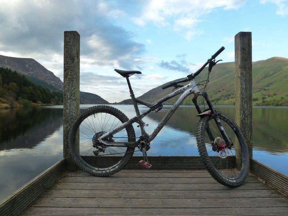 Commencal Meta AM V4: A mountain bike parked on a wooden dock next to a serene lake, surrounded by rolling hills and trees, with a cloudy sky reflecting in the water.