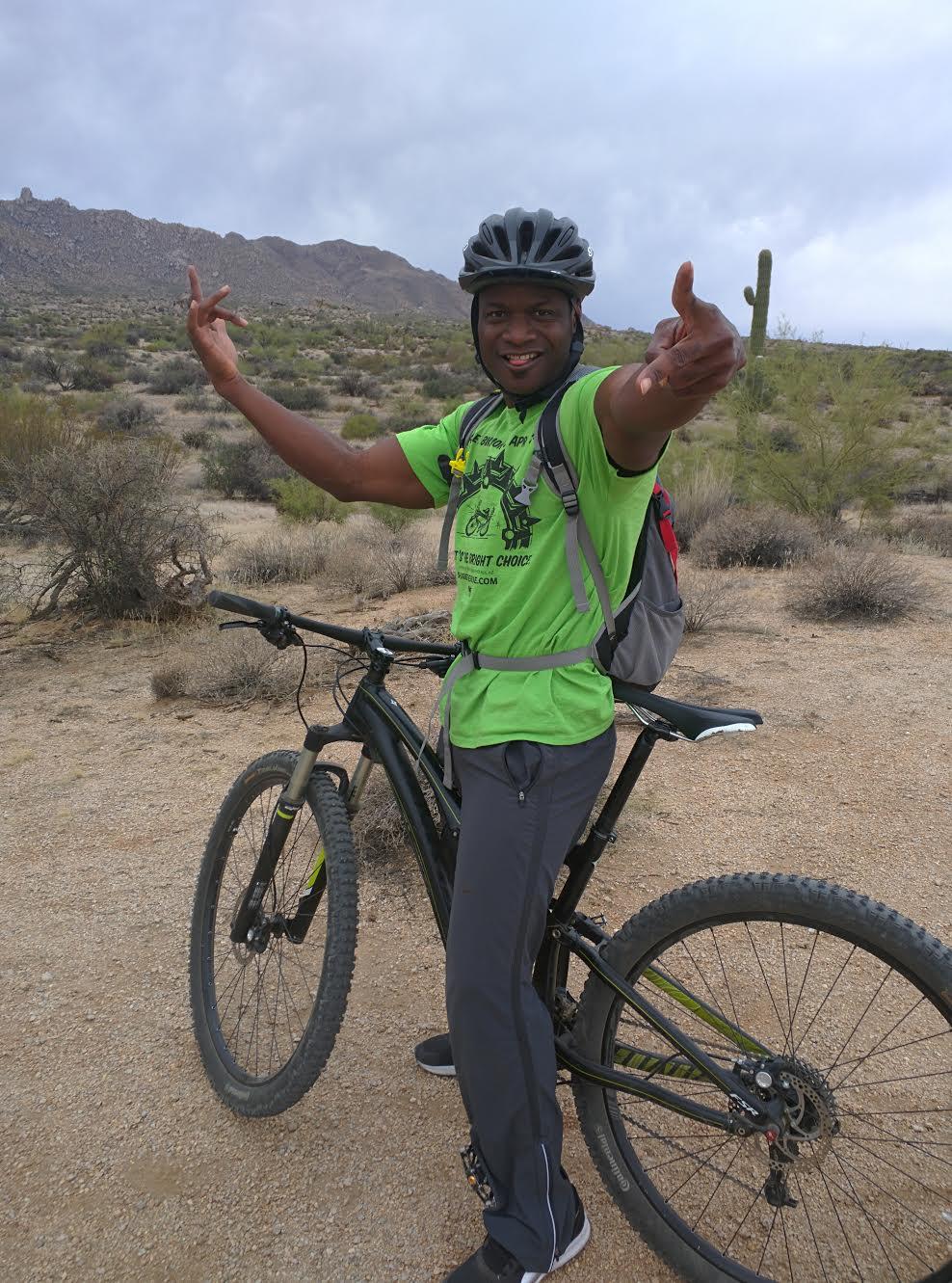 A person in a bright green t-shirt and black pants stands next to a mountain bike in a desert landscape, smiling and posing with a playful gesture. The background features rocky mountains and sparse vegetation under a cloudy sky. Pemberton Loop mountain bike trail.