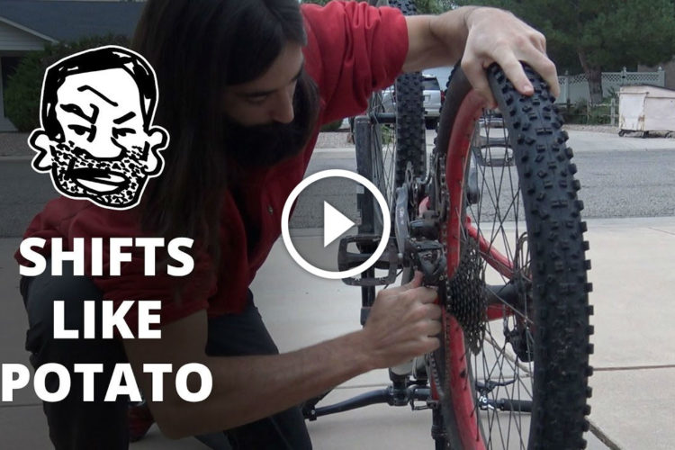 A person with long hair and a beard is crouched down, working on a bicycle's gears in a driveway. The scene includes a playful graphic of a person's face with a beard and text reading "SHIFTS LIKE POTATO." Background includes a suburban setting with trees and a house.