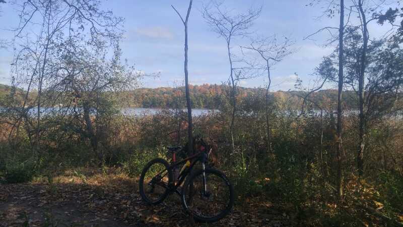 A bicycle resting on a wooded trail with a view of a lake in the background, surrounded by autumn foliage and trees under a clear blue sky. DTE Energy Foundation Trail mountain bike trail.