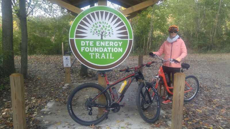 A person in a pink jacket and helmet stands beside two bicycles at the entrance of the DTE Energy Foundation Trail, marked by a large sign featuring a tree logo. Surrounding the area are trees and natural foliage, indicating a scenic outdoor setting. DTE Energy Foundation Trail mountain bike trail.