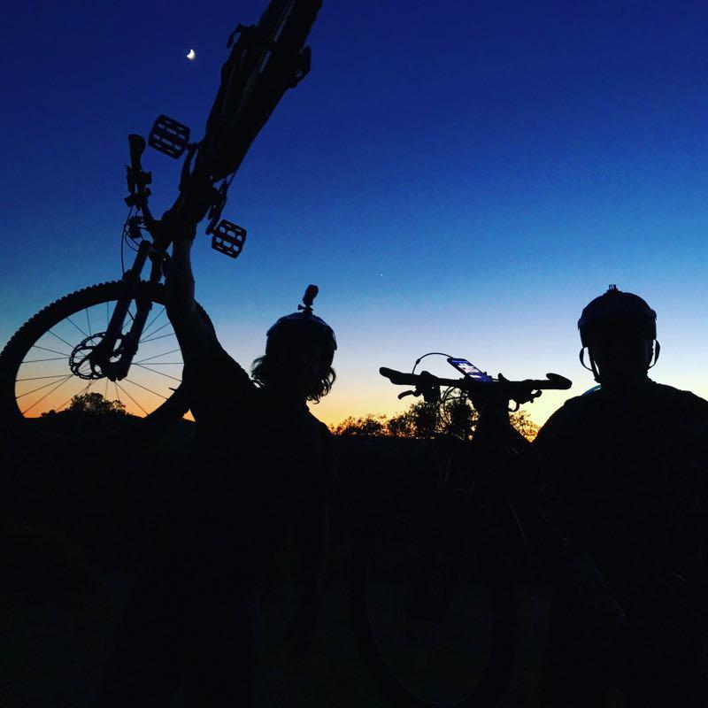 Silhouette of two mountain bikers holding their bikes aloft against a twilight sky, with a waxing crescent moon visible and warm colors of sunset in the background. Both riders are wearing helmets, and one has a camera mounted on their helmet. Annadel State Park mountain bike trail.