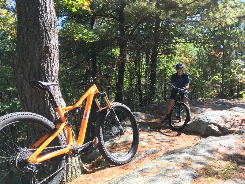 A mountain bike leaning against a tree in a forested area, with a rider sitting on a nearby rock while holding another bike. The scene is surrounded by greenery and rocky terrain, showcasing a sunny day outdoors. Vaughn Hill/bower Spring Trail System(s) mountain bike trail.