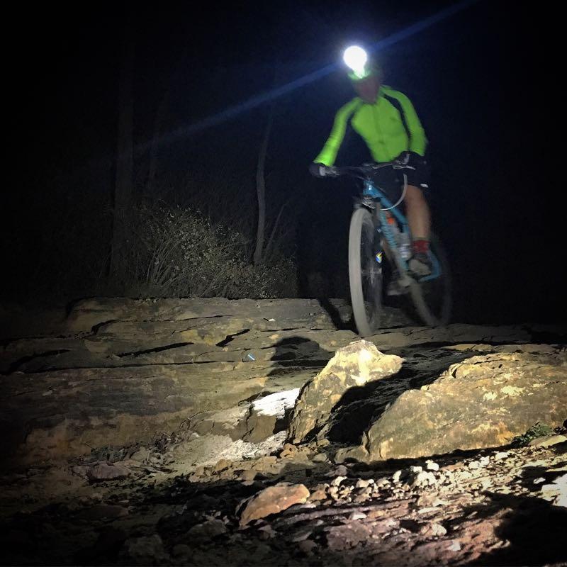 A mountain biker wearing a bright yellow jacket and headlamp rides over rocky terrain at night, with trees and dark surroundings visible in the background. Quarry Ridge Park mountain bike trail.
