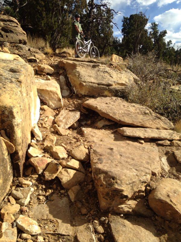 A person standing next to a mountain bike on rocky terrain, surrounded by sparse vegetation and trees under a partly cloudy sky. The path is made up of large, uneven stones, indicating a rugged outdoor environment. Oil Well Flats mountain bike trail.