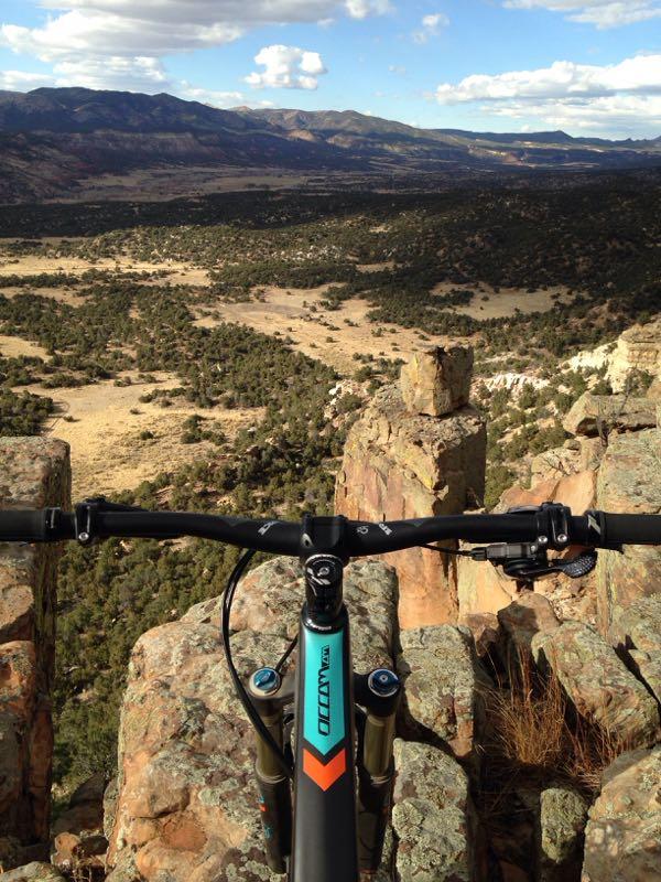 A mountain bike positioned near the edge of a rocky cliff, overlooking a vast landscape of rolling hills, trees, and distant mountains under a partly cloudy sky. Oil Well Flats mountain bike trail.