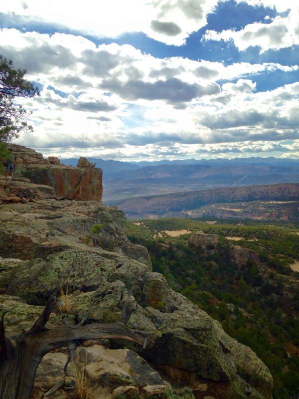 Scenic view from a rocky cliff, showcasing layered mountains and a vast landscape under a partly cloudy sky. The foreground features textured rock formations and sparse vegetation, while the background displays rolling hills and distant peaks. Oil Well Flats mountain bike trail.