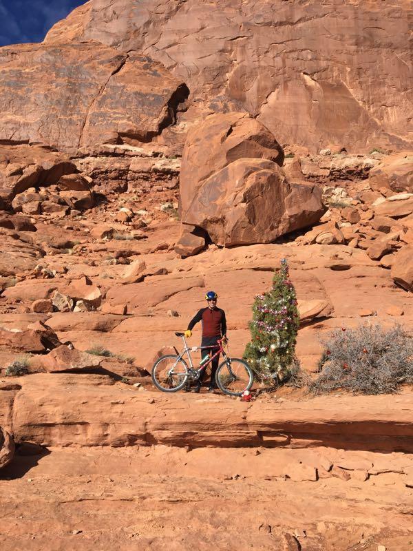A cyclist stands next to a decorated Christmas tree on a rocky desert landscape, with large red rock formations in the background. The cyclist is wearing a helmet and has a mountain bike beside them. The scene captures a blend of holiday spirit and rugged outdoor adventure. Navajo Rocks mountain bike trail.