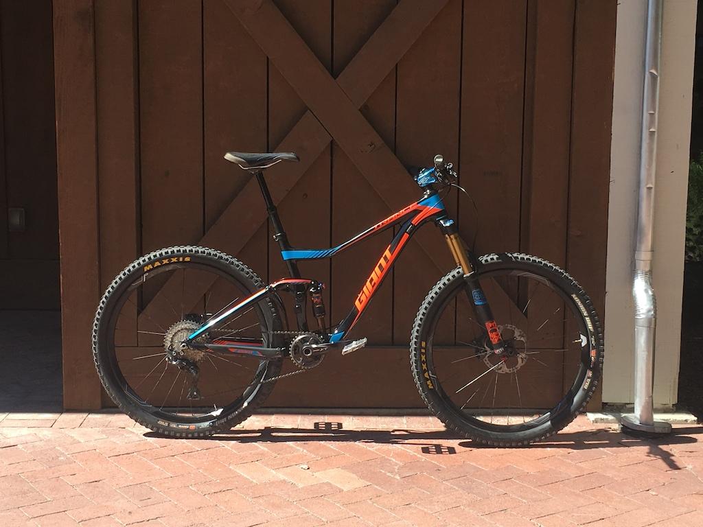 Giant Trance Advanced 1 27.5: A brightly colored mountain bike with an orange and blue frame, featuring a front suspension fork and knobby tires, leaning against a wooden wall with a rustic design. The bike is positioned on a brick surface, showcasing its detailed components and sturdy build.