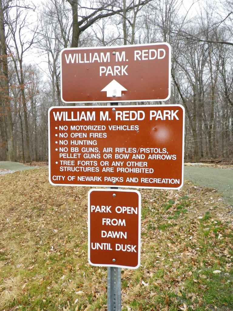 A brown informational sign for William M. Redd Park, displaying rules and regulations. The top section indicates the park's name, while the lower section lists prohibitions such as no motorized vehicles, open fires, or hunting, and states that the park is open from dawn until dusk. The background features a wooded area with bare trees. Redd Park mountain bike trail.