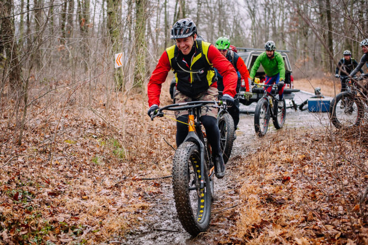 A group of four mountain bikers riding on a muddy trail through a forest in autumn. The lead rider is wearing a red long-sleeve shirt, a black vest, and a helmet, smiling as he navigates the path. The other bikers are following closely behind, dressed in colorful cycling gear. Trees with bare branches and fallen leaves cover the ground, creating a natural setting. A vehicle is parked in the background.