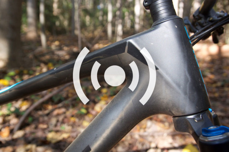 Close-up view of a mountain bike frame, featuring a sleek design with a dark finish and blue accents. The background is a natural setting with soft focus showing trees and fallen leaves. In the center, a symbol indicating connectivity or signal is overlaid, suggesting potential smart features of the bike.