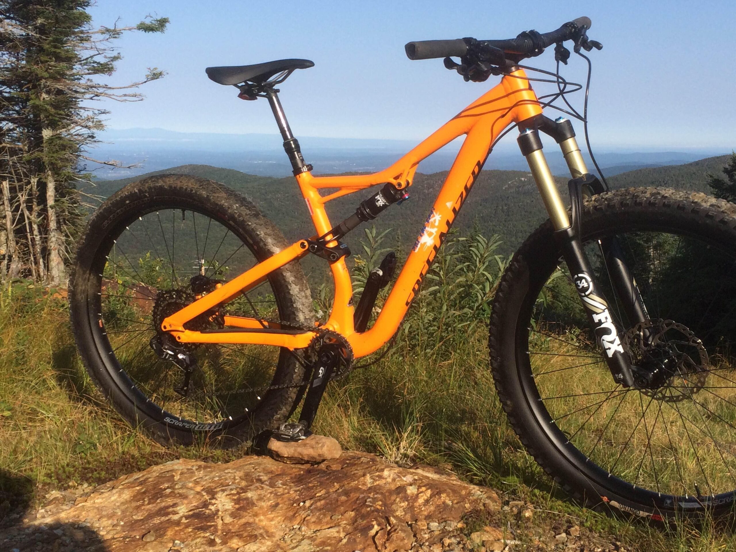 Specialized Stumpjumper FSR 6Fattie: An orange mountain bike is positioned on a large rock, with a scenic view of green hills and a clear blue sky in the background. The bike features a sturdy frame, thick tires, and front suspension, emphasizing its off-road capabilities. Tall grass and trees are visible in the surrounding landscape.