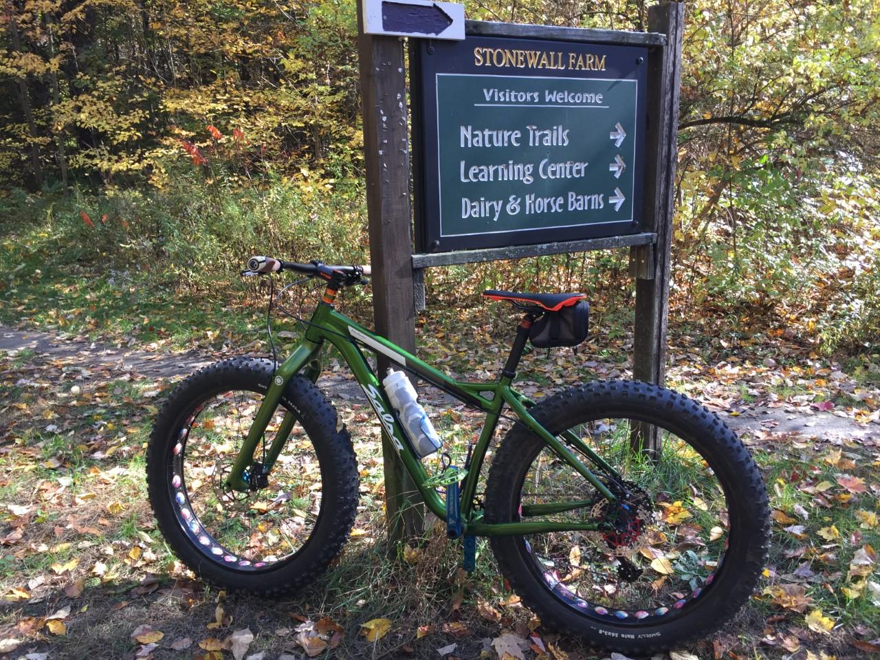 Salsa Blackborow: A green fat tire bike parked next to a sign for Stonewall Farm, with directions to nature trails, a learning center, and dairy and horse barns. The scene features colorful autumn foliage and fallen leaves on the ground.