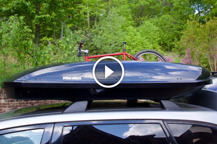 A roof-mounted cargo box by Yakima is secured on the roof of a silver car, with a red mountain bike resting on a bike rack. The background features lush green trees and blooming flowers.
