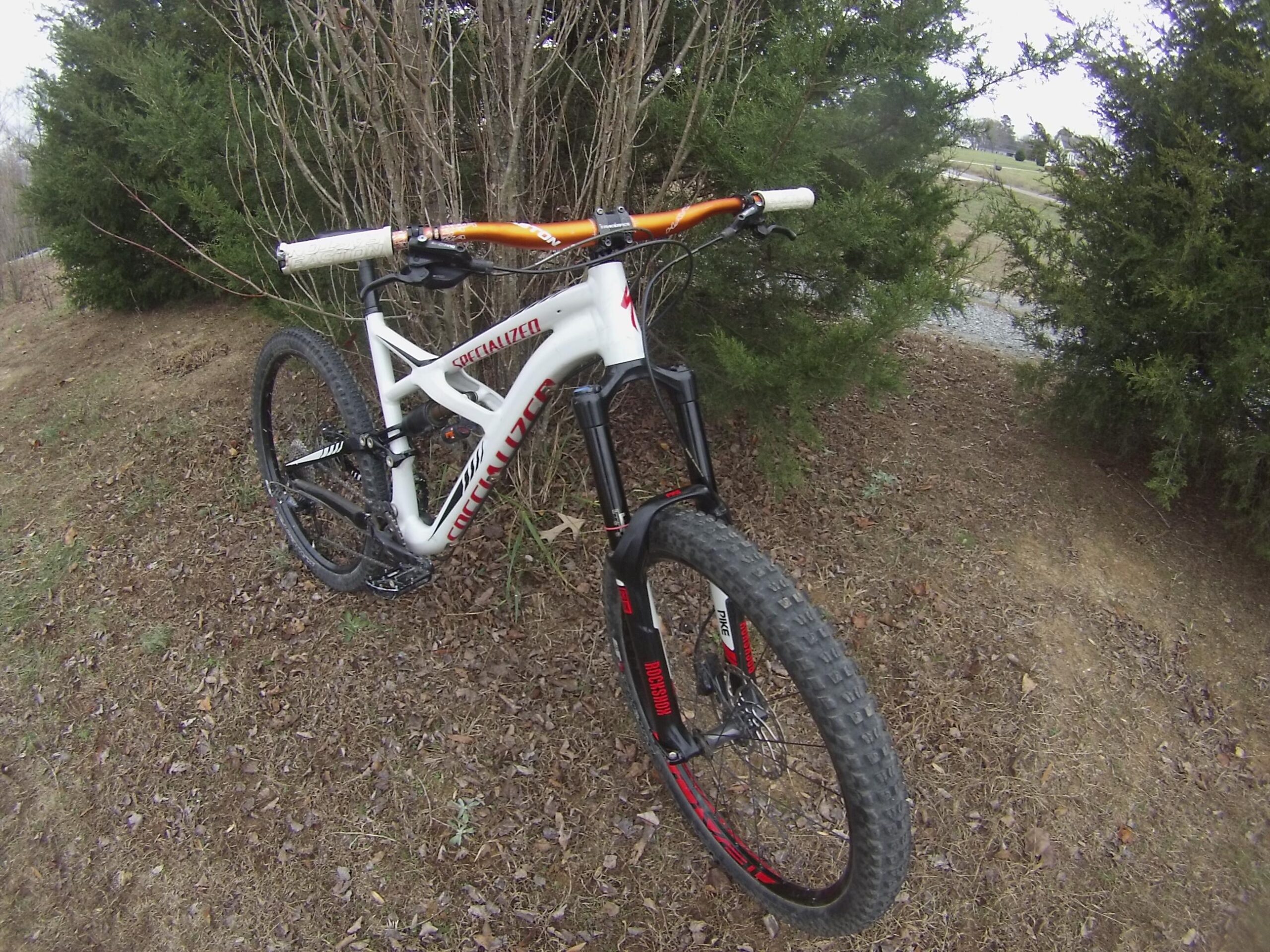 Specialized Enduro Comp 650b: A close-up view of a mountain bike positioned on a dirt path, surrounded by shrubs. The bike features a white frame with red branding, an orange handlebar, and thick knobby tires suitable for off-road riding. The background includes a blurred view of grass and trees, suggesting an outdoor setting.