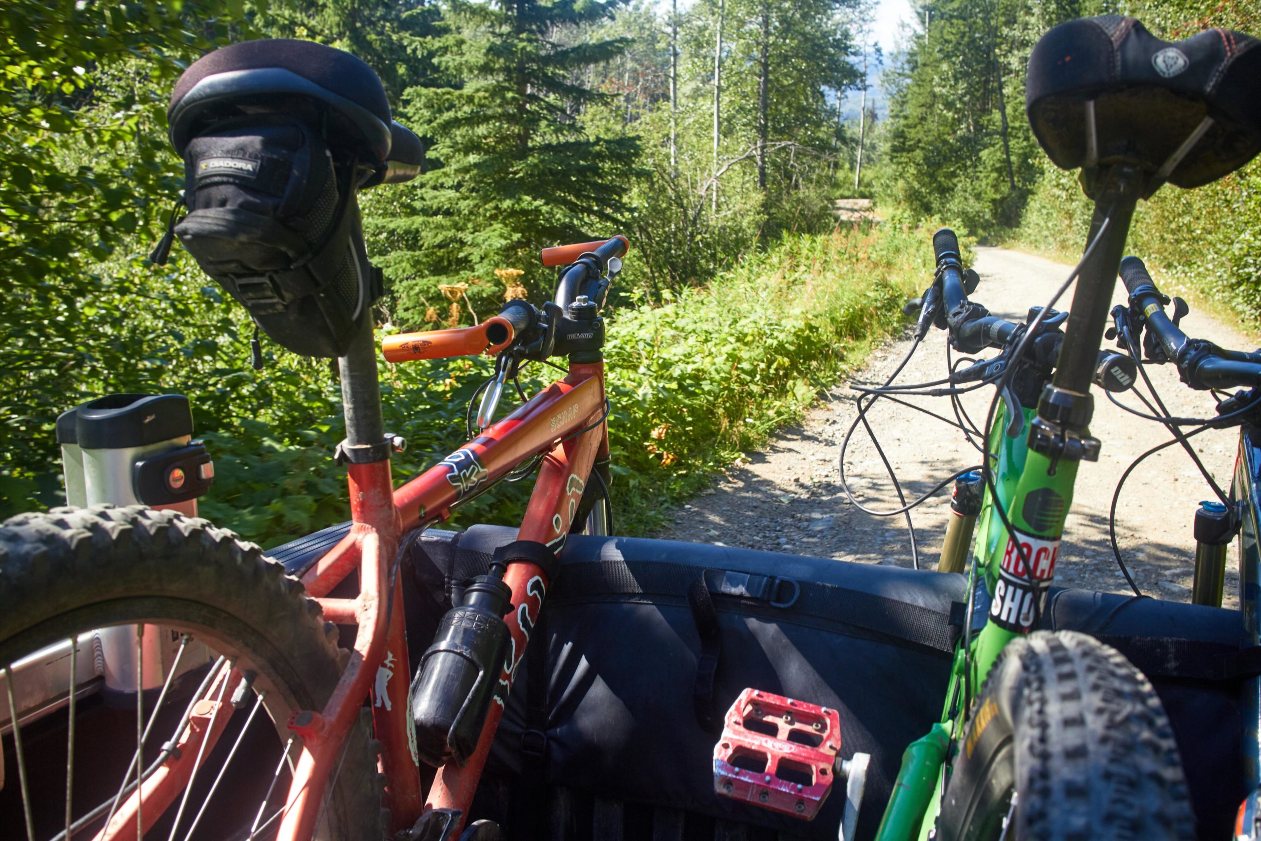 Two bicycles, one red and one green, are positioned in the back of a vehicle on a gravel path surrounded by trees and greenery. A water bottle and a pedal can be seen next to the bikes. The scene suggests an outdoor adventure or a mountain biking trip. Boer Mountain mountain bike trail.