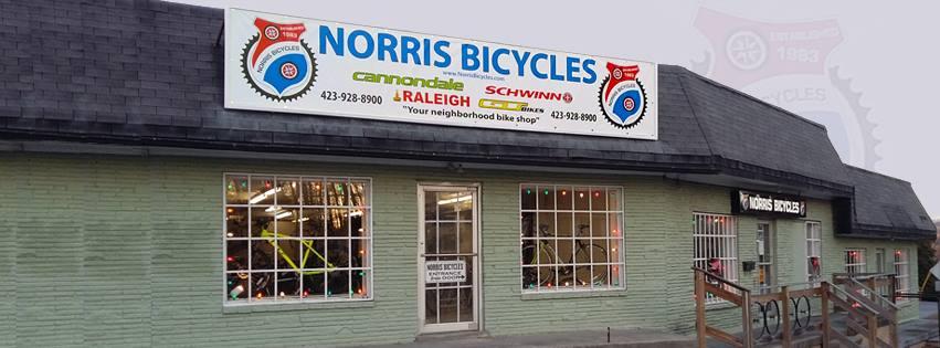 Image of the exterior of Norris Bicycles, featuring a sign with the store name and bicycle brand logos, including Cannondale, Raleigh, and Schwinn. The building has large windows displaying bicycles inside and a welcoming entrance with steps leading up to the door. The sign emphasizes the store