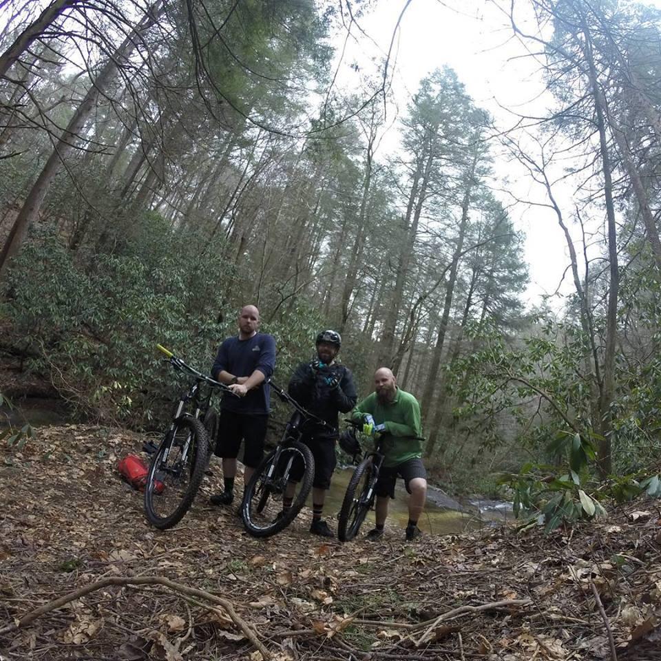 Three mountain bikers pause on a forest trail next to a stream. They are standing beside their bikes, surrounded by trees and fallen leaves. One biker wears a helmet and a jacket, while the others are dressed in casual biking attire. The scene captures the natural beauty of the woods, emphasizing an outdoor adventure. Bull / Jake Mountain mountain bike trail.