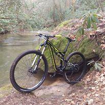 A mountain bike leaning against a large rock near a flowing stream, surrounded by lush greenery and trees in a serene outdoor setting. Bull / Jake Mountain mountain bike trail.