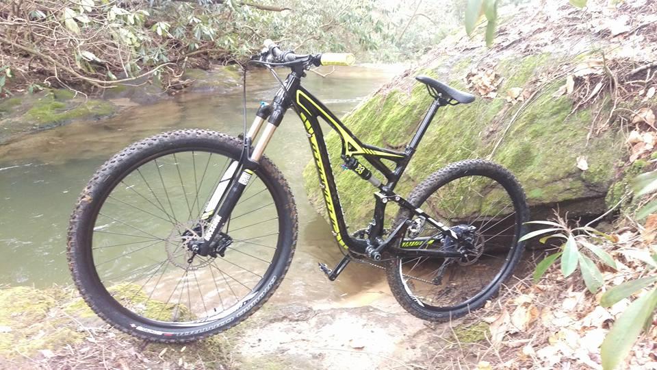 A mountain bike rests against a moss-covered rock near a flowing stream, surrounded by lush greenery and fallen leaves. The bike features a sleek black frame with yellow accents and rugged tires, suggesting it is ready for off-road adventures. Bull / Jake Mountain mountain bike trail.