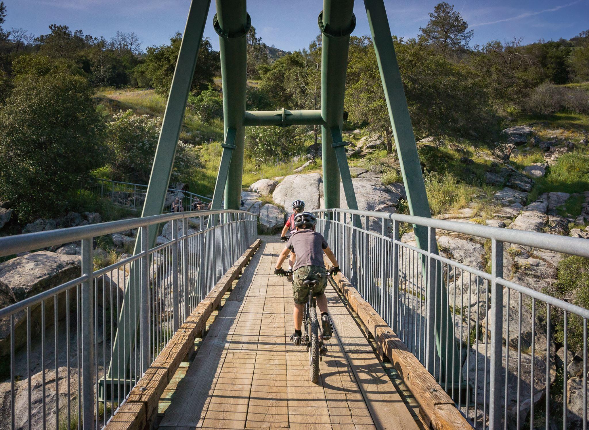 Two cyclists ride across a wooden bridge surrounded by greenery and rocky terrain. The bridge features green metal supports and safety railings, with sunlight illuminating the scene. San Joaquin River Trail mountain bike trail.