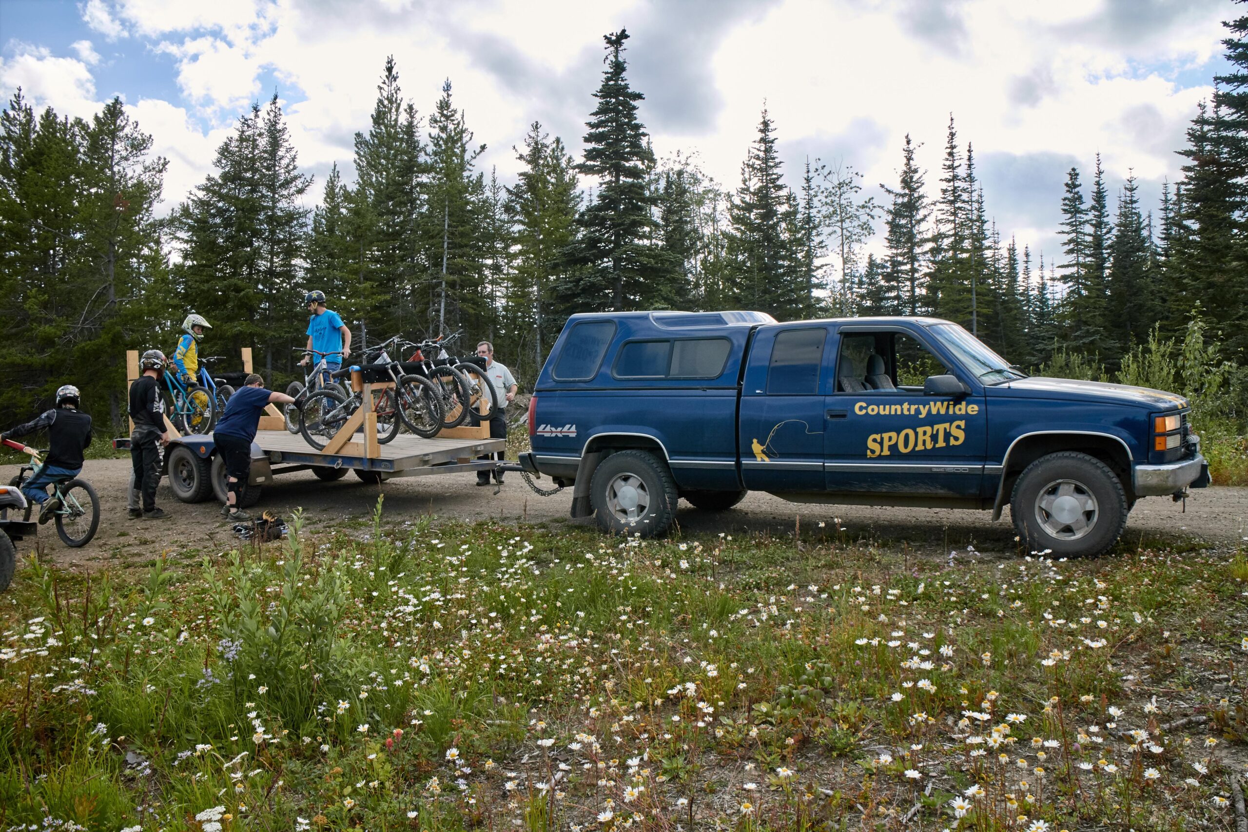 A blue truck with “CountryWide Sports” branding parked next to a trailer loaded with several mountain bikes. People are actively unloading bikes from the trailer in a forested area surrounded by wildflowers. The sky is partly cloudy, and tall evergreen trees are visible in the background.