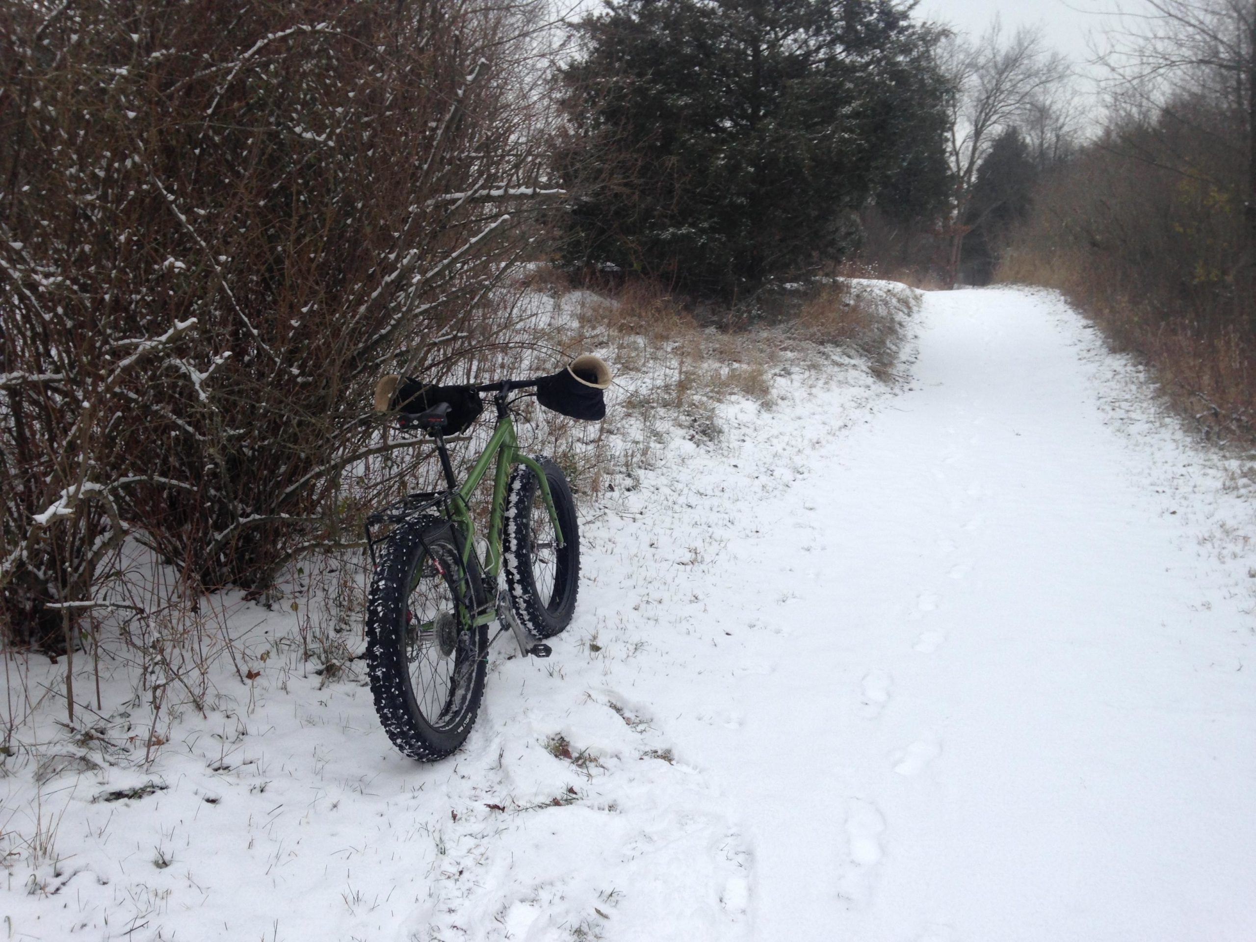 A green fat-tire bicycle parked beside a snow-covered trail, surrounded by bare bushes and trees. The ground is blanketed in fresh snow, and footprints are visible in the snow leading down the path. The scene captures a quiet winter landscape. Potawatomi trail mountain bike trail.