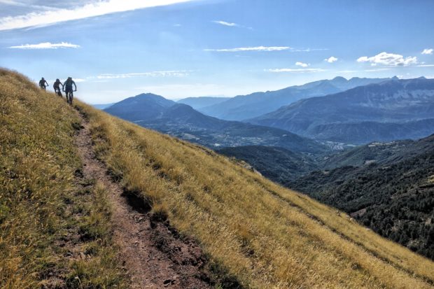 Three mountain bikers navigate a dirt trail along a grassy hillside, overlooking a scenic valley and distant mountains under a bright blue sky with scattered clouds.