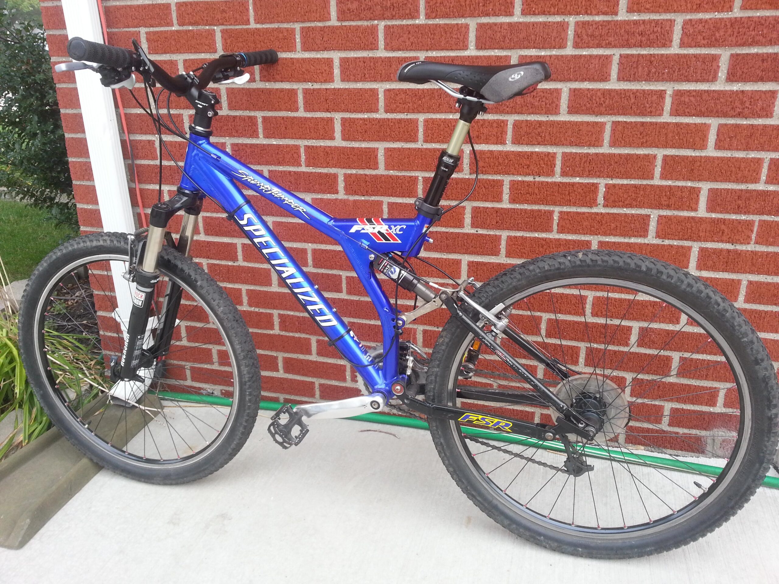 Specialized Stumpjumper FSR: A blue mountain bike leanings against a red brick wall, featuring a front suspension fork, textured tires, and a black seat. The bike's frame displays the "Specialized" logo prominently.