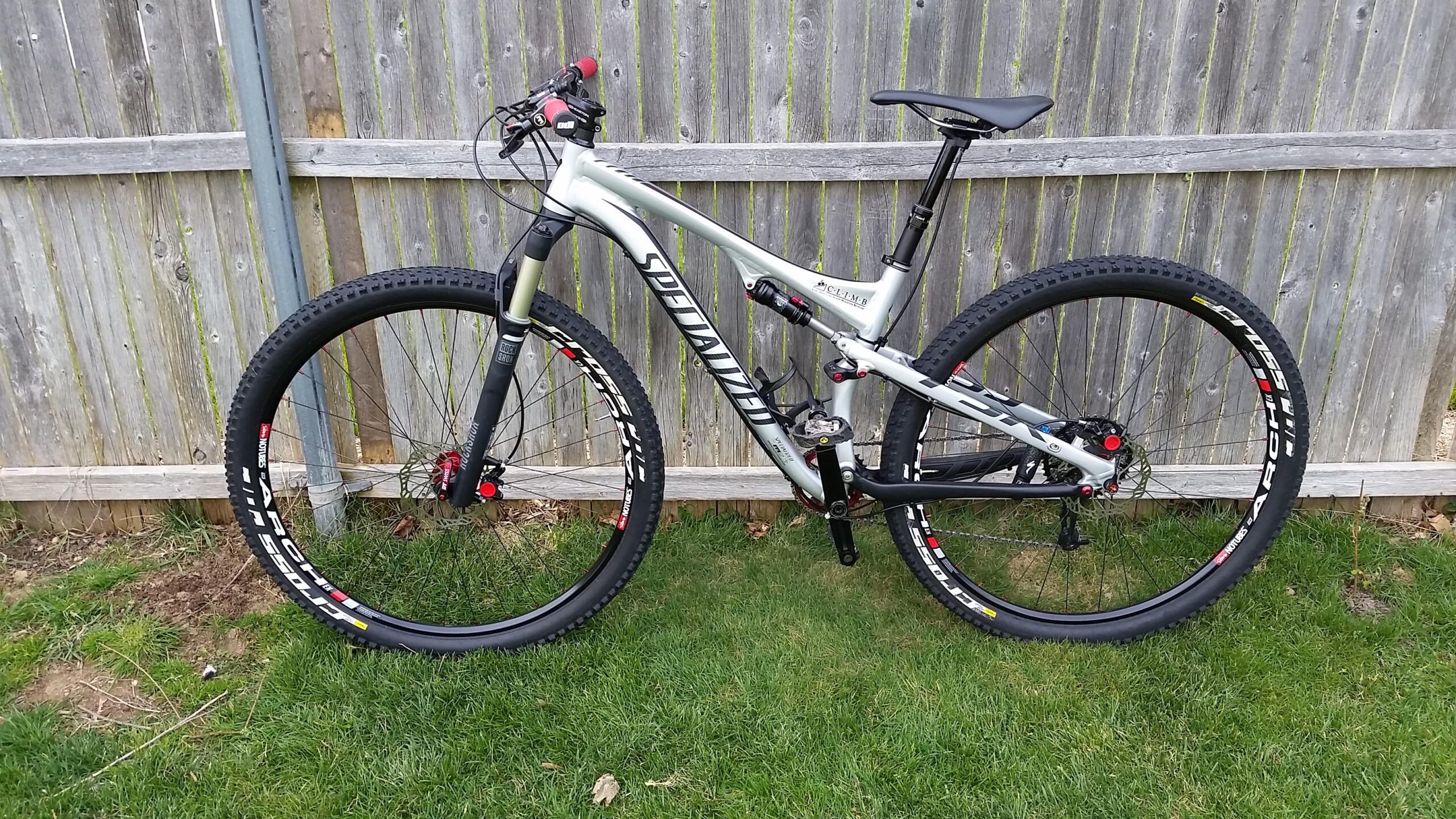 Specialized Epic Comp: A silver and black mountain bike stands on grass beside a wooden fence. The bike features thick tires, front suspension, and a sleek frame design, with components visible such as the handlebars and seat.