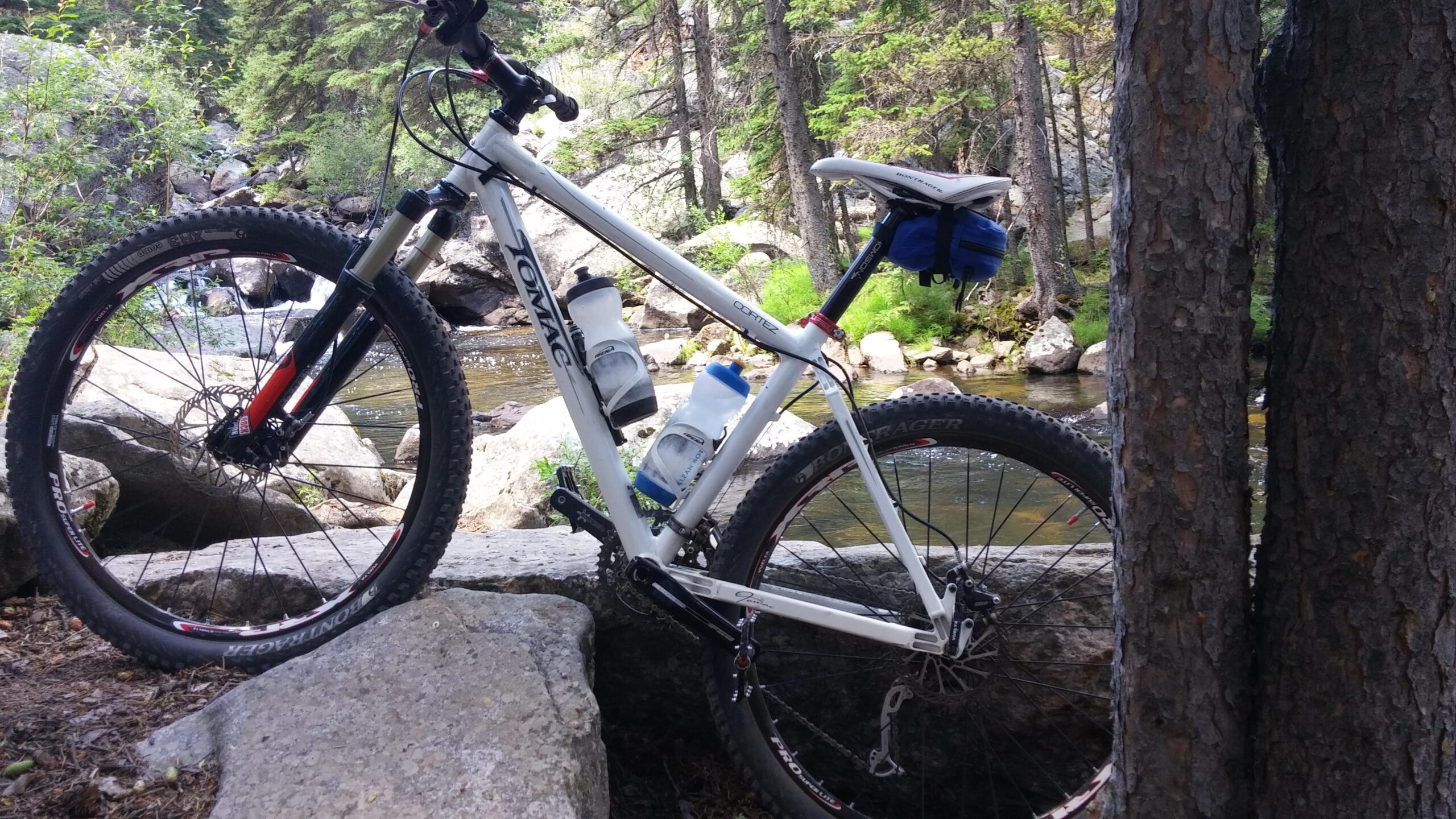 Tomac Cortez: A white mountain bike with two water bottles attached is leaning against a large rock by a stream in a forested area. The background features lush greenery and flowing water, suggesting a peaceful outdoor setting ideal for cycling and nature exploration.