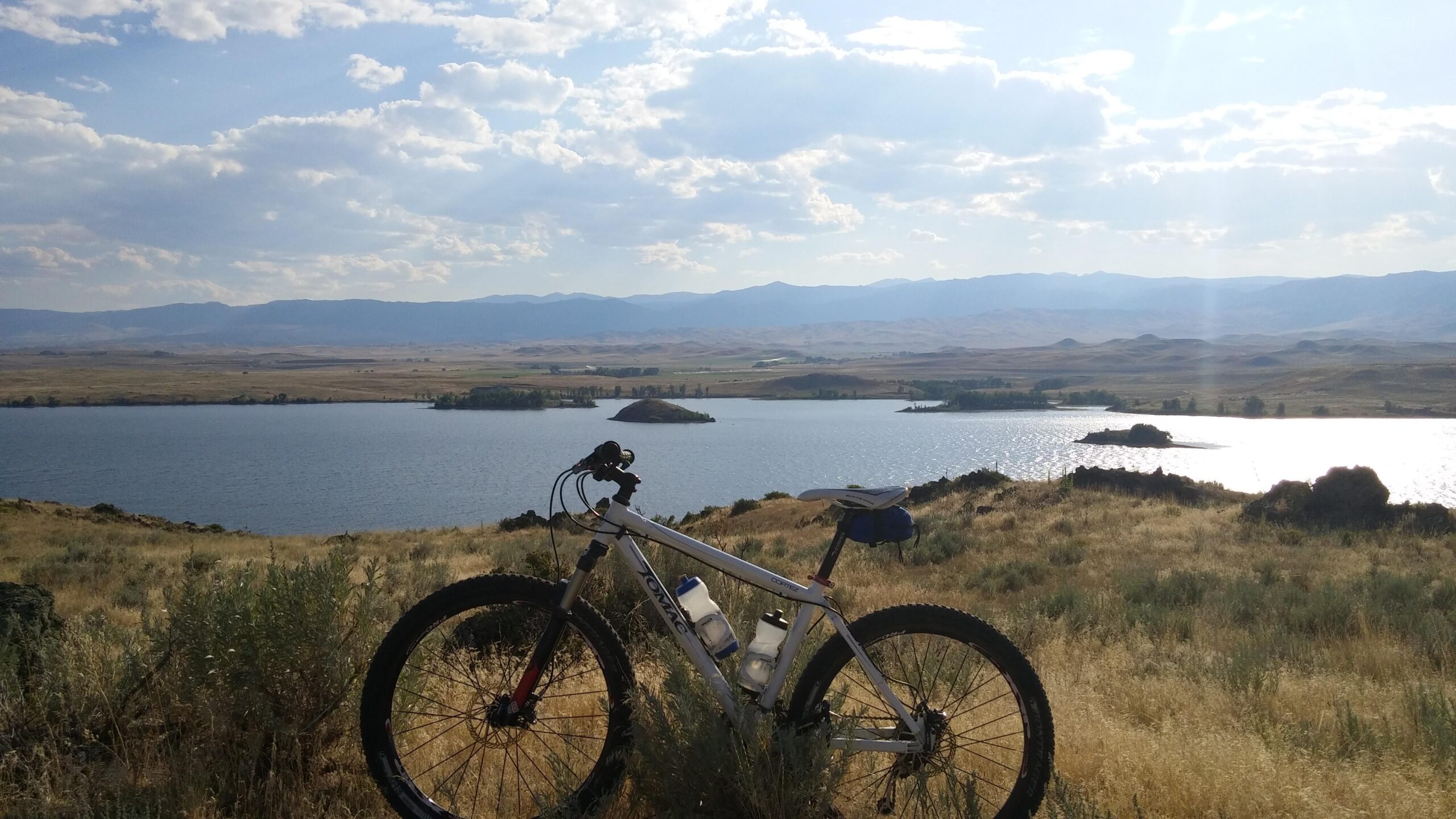 Tomac Cortez: A mountain bike resting on a grassy hillside overlooking a calm lake, with distant mountains and a partly cloudy sky in the background.