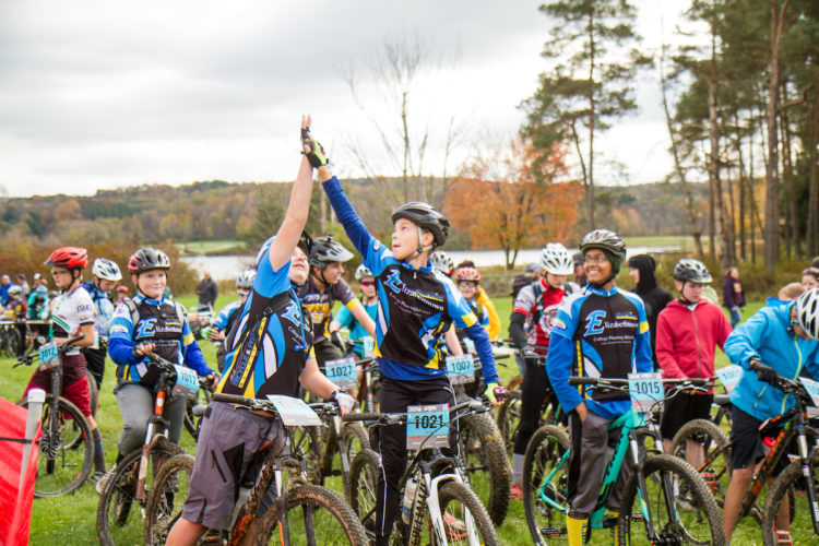 A group of young cyclists in colorful jerseys and helmets gather for a race, with some participants high-fiving and showing excitement. The background features a scenic view of trees and a lake, suggesting an outdoor event on an overcast day.
