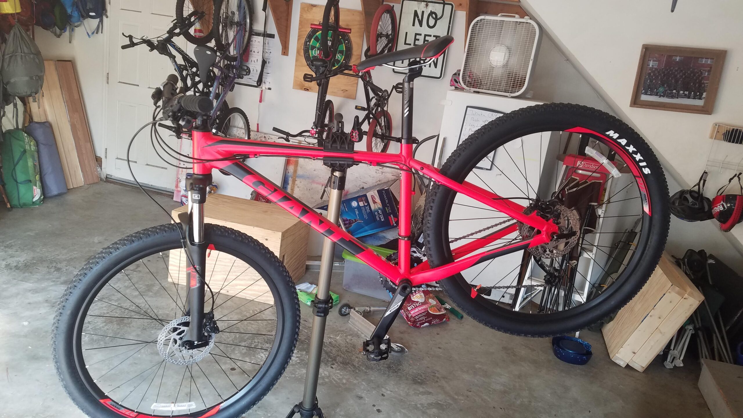 Giant Talon 27.5 2: A bright red mountain bike is mounted on a repair stand in a garage. The bike features thick tires and a disc brake system. In the background, there are bicycles hung on the wall, various tools, a fan, and a wooden box. The garage has a concrete floor and some items including a rug and a blue bowl are visible.