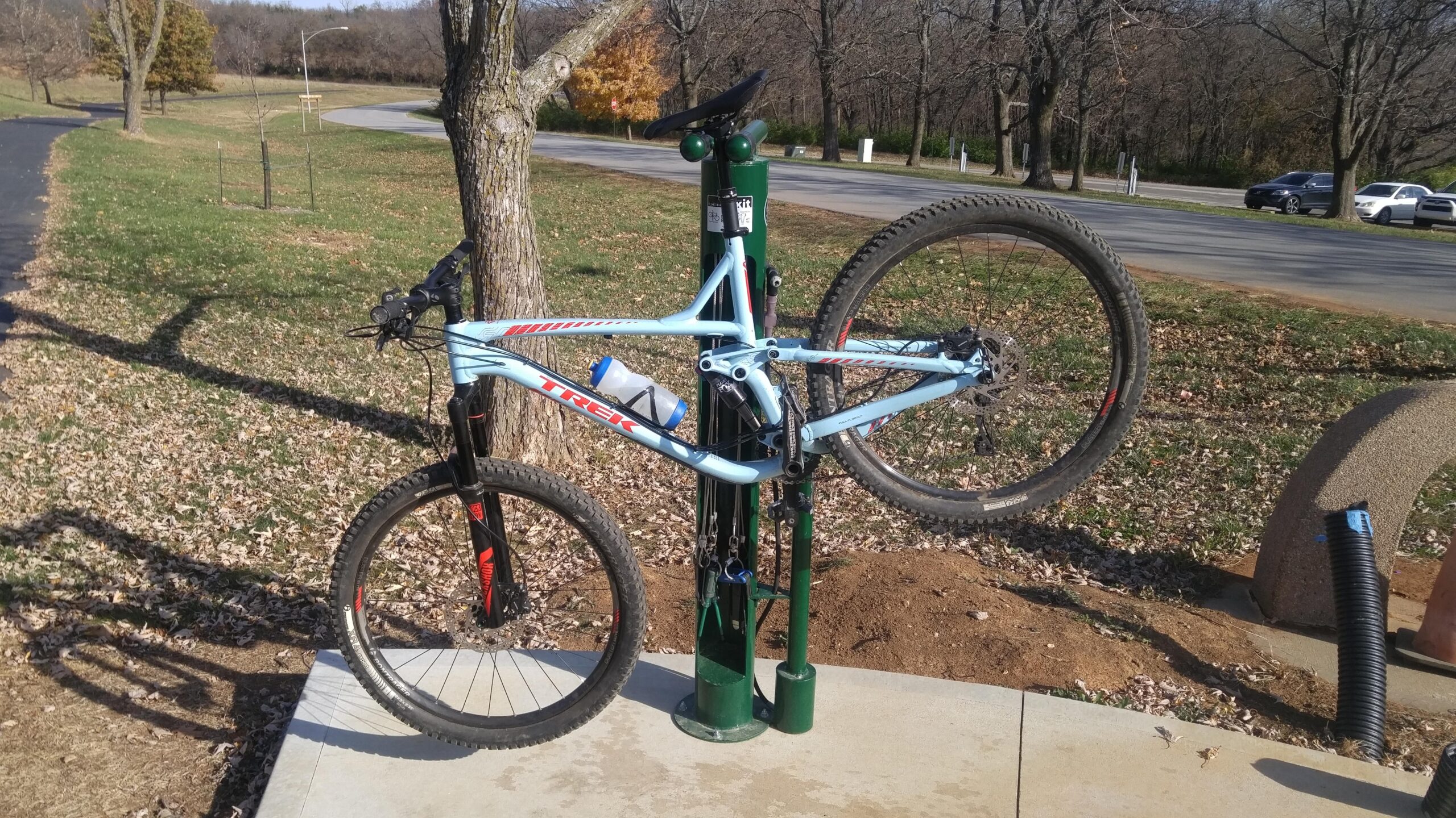 Trek trek remedy 7 27.5: A light blue bicycle is mounted on a bike repair stand in a park setting. The bike's front wheel is lifted off the ground, displaying its tires and frame. Nearby, there are fallen leaves on the ground, a tree in the background, and a paved path leading to the road. Tools and equipment are attached to the bike stand for maintenance purposes.
