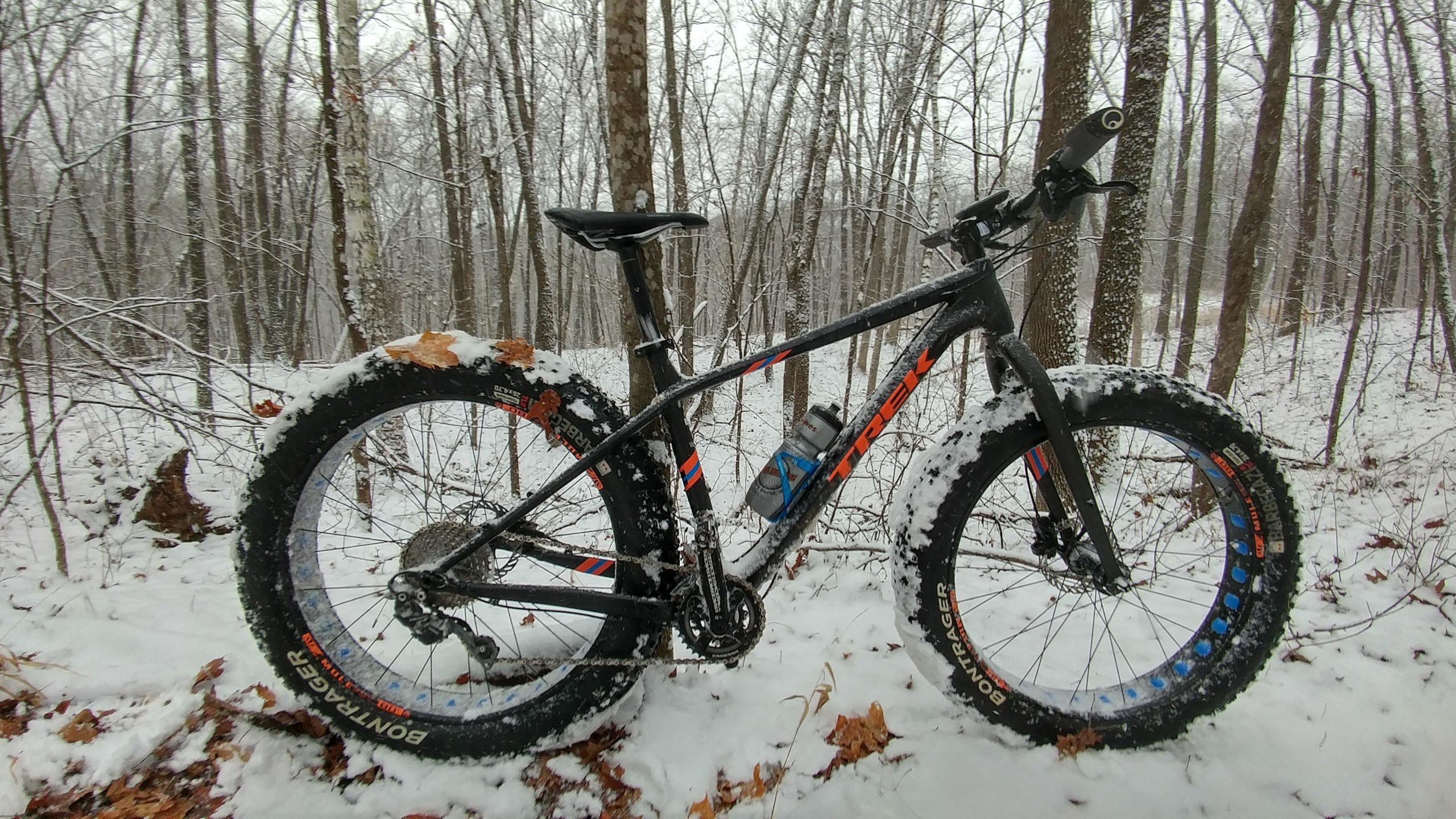 A fat tire mountain bike stands on a snowy forest path, surrounded by bare trees and a light snowfall. The bike features a black frame with orange and blue accents, and its large tires are partly covered in snow, emphasizing its winter-ready design. Greenbush mountain bike trail.