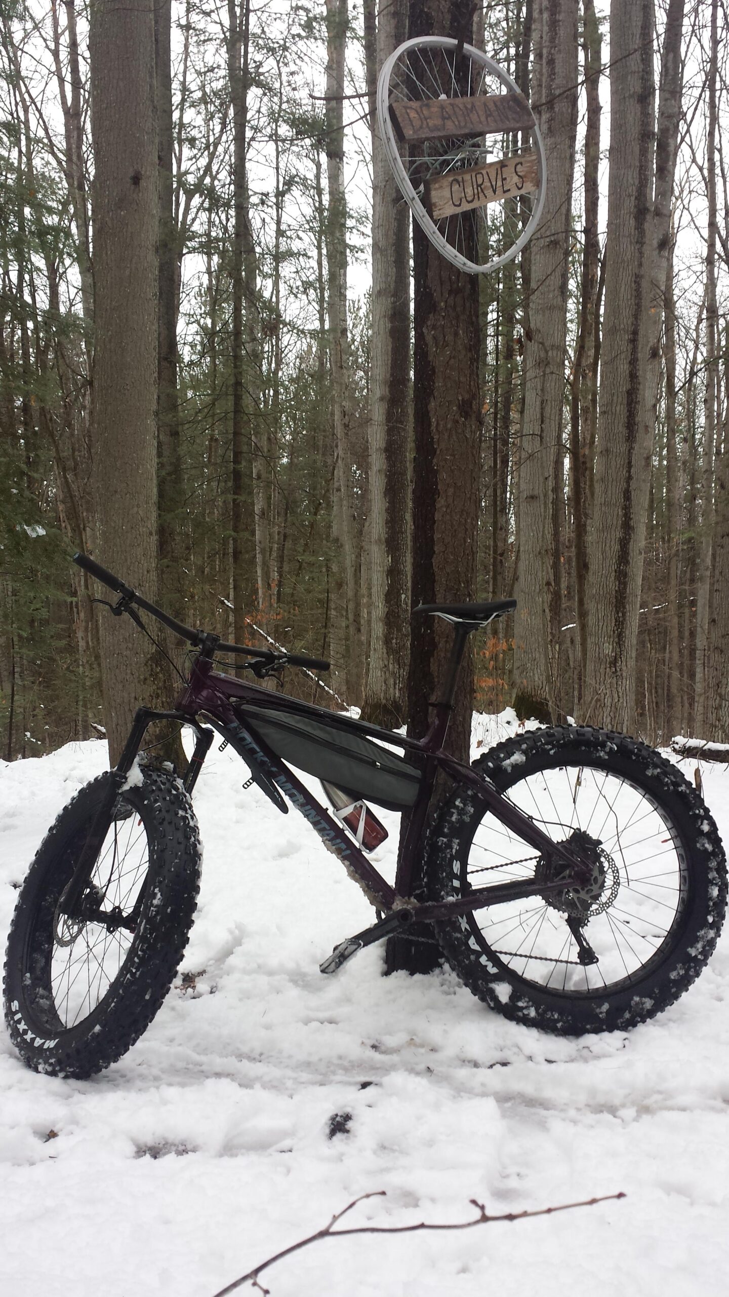A fat tire mountain bike rests on a snowy trail in a forest, with tall trees in the background. A wooden sign reading "Deadman's Curves" is attached to a tree, framed by a bicycle rim. Snow covers the ground, creating a wintery backdrop for the bike.
