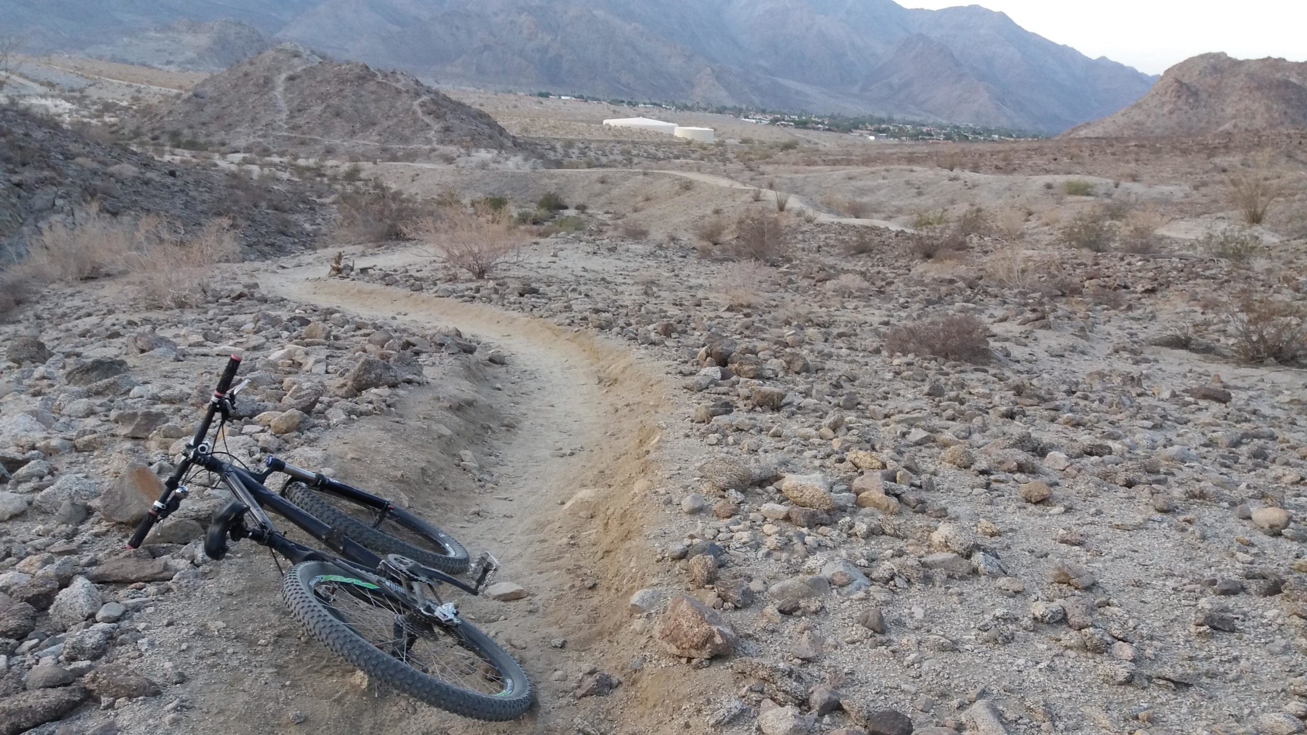 A mountain bike resting on its side beside a rocky, winding trail in a desert landscape, with hills and a distant settlement in the background under a cloudy sky. The Cove to Lake Trail mountain bike trail.