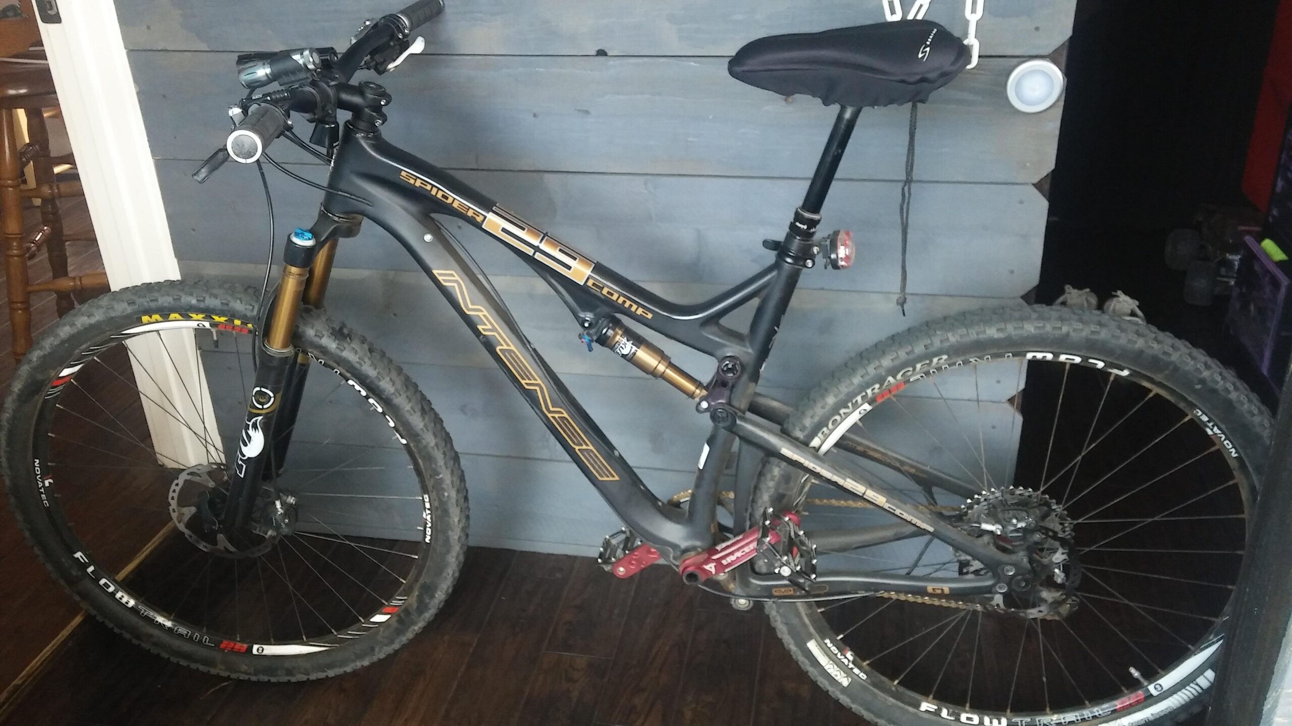 Intense Spider 29 Comp: A black mountain bike with gold and red accents resting against a gray wooden wall. The bike features a suspension fork, disc brakes, and knobby tires suitable for off-road riding. There is a saddle on the seat post and various components visible, such as the handlebars and gear shifters. The background includes part of a chair and a dimly lit area.